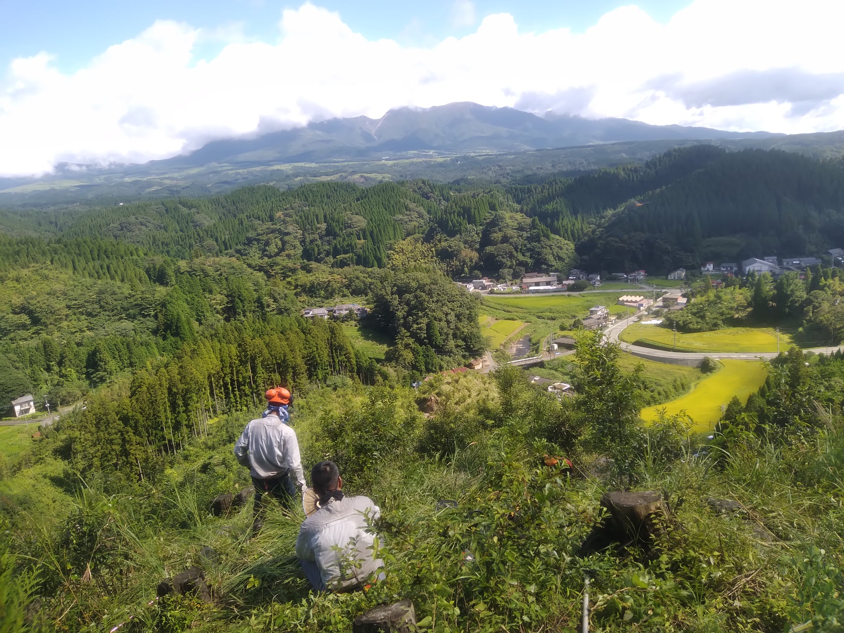 山仕事の合間に久住山と村を見渡す男性作業員2名
